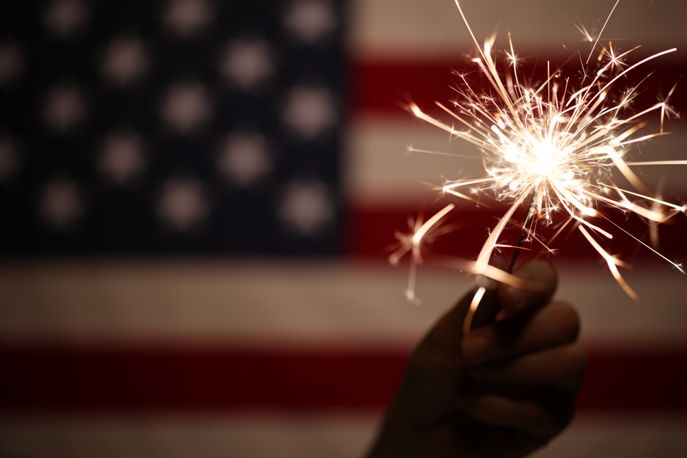 A hand holding a lit sparkler in the foreground, with a blurred American flag in the background.